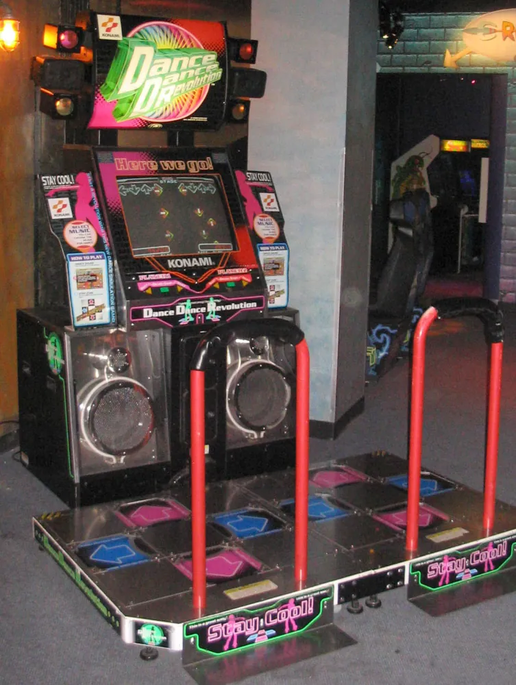 A Dance Dance Revolution arcade machine. It has a CRT monitor and two metal platforms with four buttons for players to step on.
